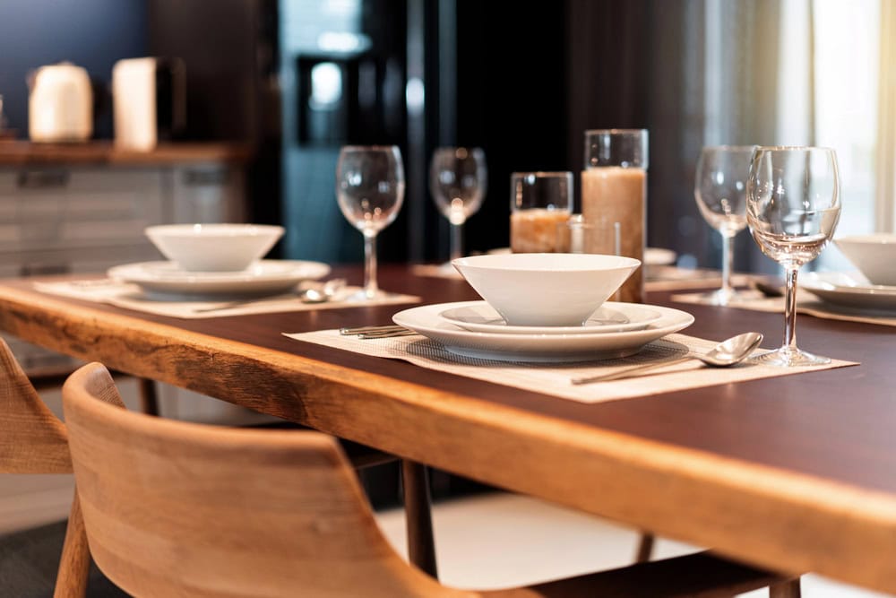A wooden dining table set with white plates, bowls, wine glasses, and silverware on placemats, with three candles and wooden chairs around it.