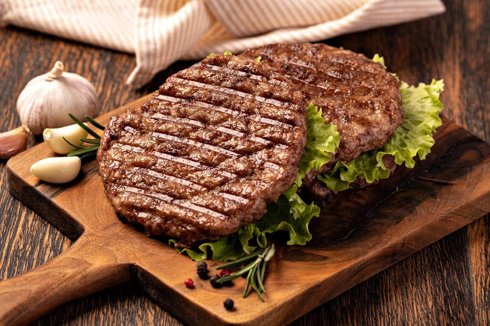 Two grilled beef burger patties on a wooden board, served on lettuce leaves with garlic, peppercorns, and rosemary beside them.