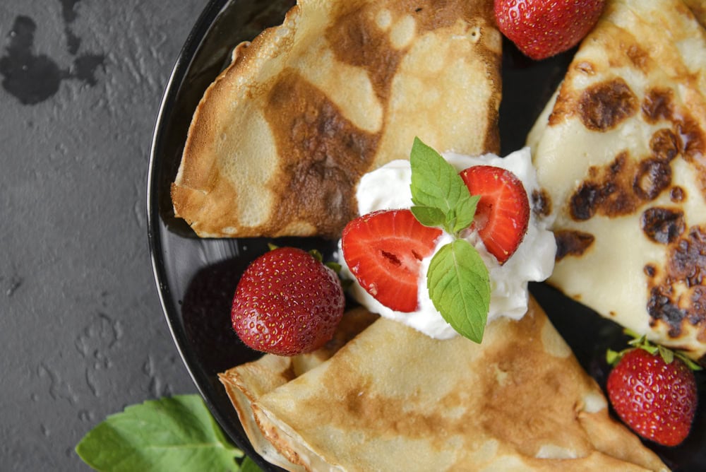 A plate with folded crepes, fresh strawberries, a dollop of whipped cream, and mint leaves on a dark background.
