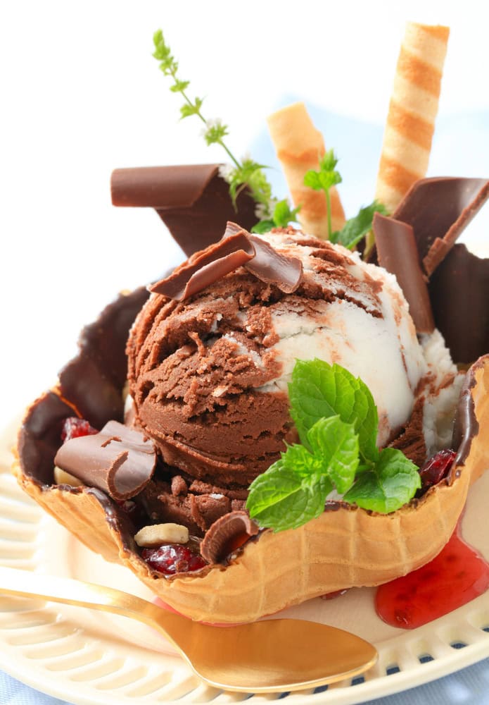 A waffle bowl filled with chocolate and vanilla ice cream, chocolate pieces, wafer rolls, mint leaves, and a red sauce garnish, served on a plate with a spoon and fork.