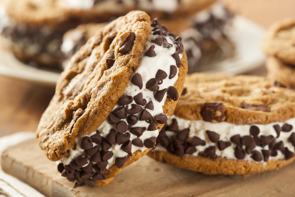 Two chocolate chip cookie ice cream sandwiches with vanilla ice cream and mini chocolate chips on the sides, placed on a wooden surface.