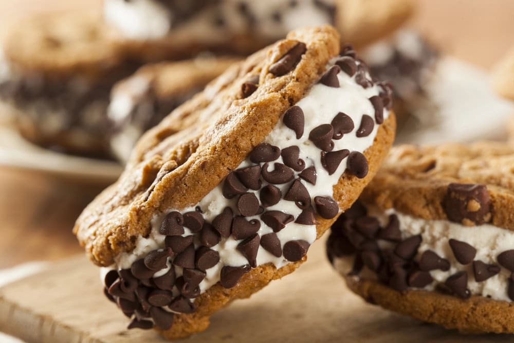 A close-up of an ice cream sandwich made with chocolate chip cookies and vanilla ice cream, with chocolate chips coating the ice cream edges.