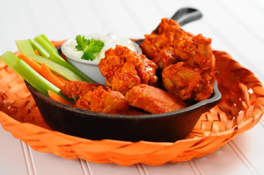 Buffalo chicken wings in a skillet with carrot and celery sticks, served with a side of ranch dip, placed in an orange woven basket.