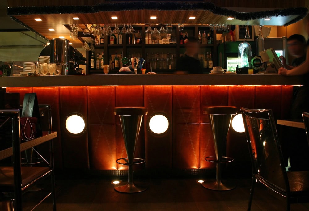 A dimly lit bar with three metal stools, illuminated panels beneath the counter, and shelves stocked with bottles and glasses in the background.