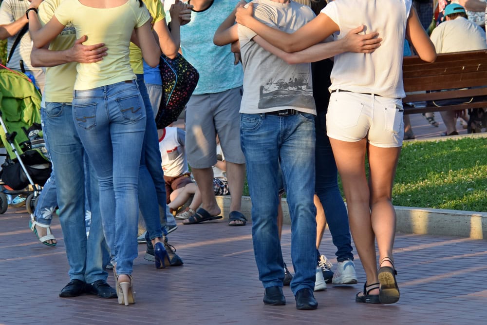 Several pairs of people dance outdoors on a paved area, with others standing and sitting on benches in the background.