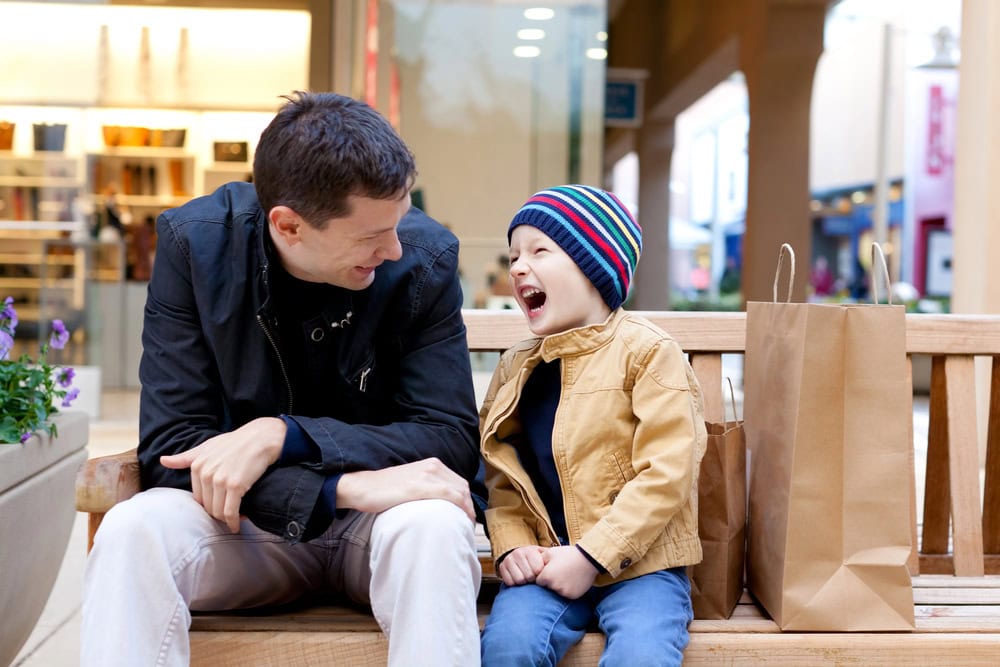 An adult and a child sit on a bench with shopping bags, smiling and laughing together in an outdoor shopping area.