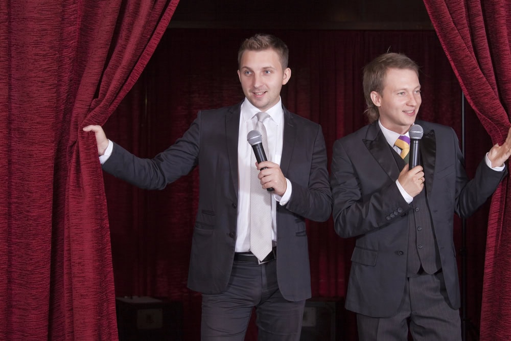 Two men in suits hold microphones while standing between red stage curtains.