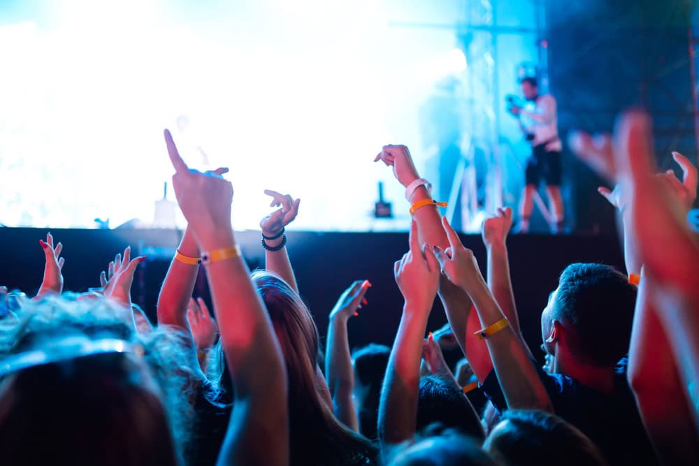 A crowd of people with raised hands at a concert, illuminated by bright stage lights, with a cameraman visible in the background.