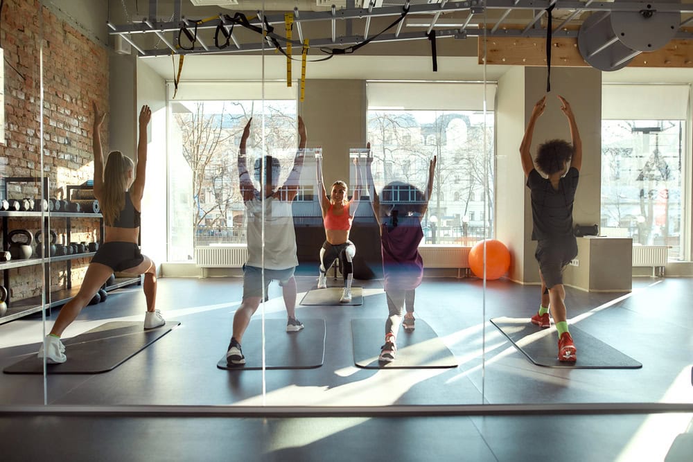 Five people participate in a fitness class, following an instructor at the front in a gym with large windows and exercise mats.