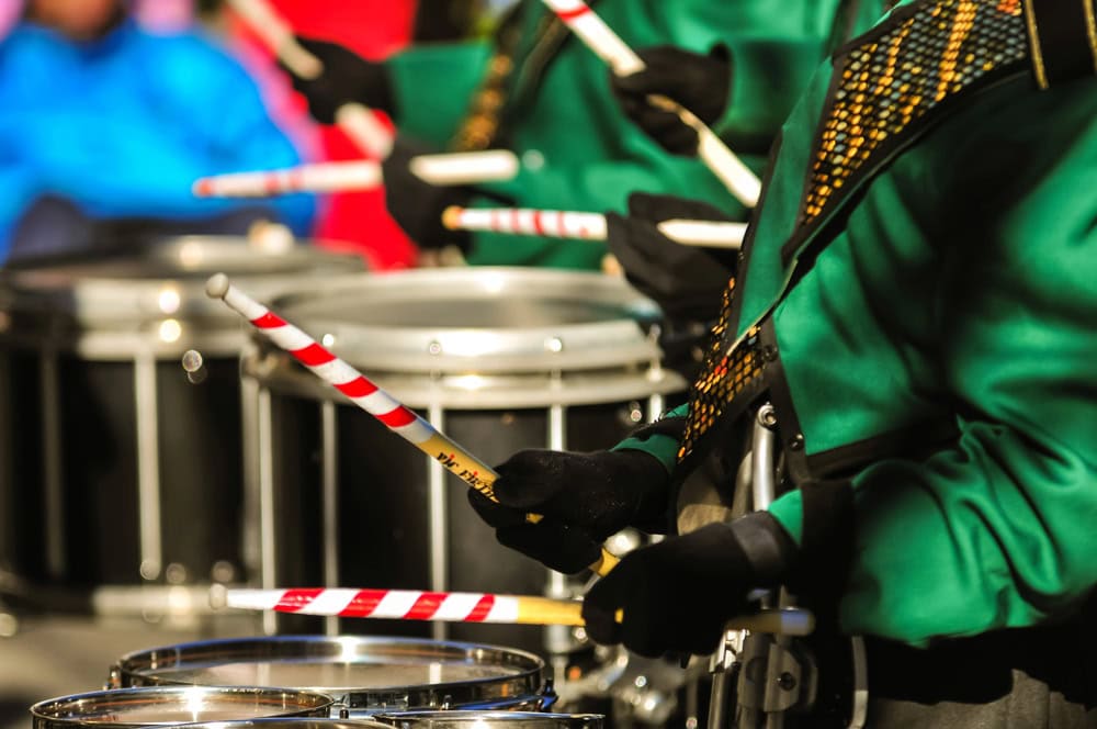 Close-up of drummers in green uniforms playing snare drums with red-and-white striped drumsticks during a performance.