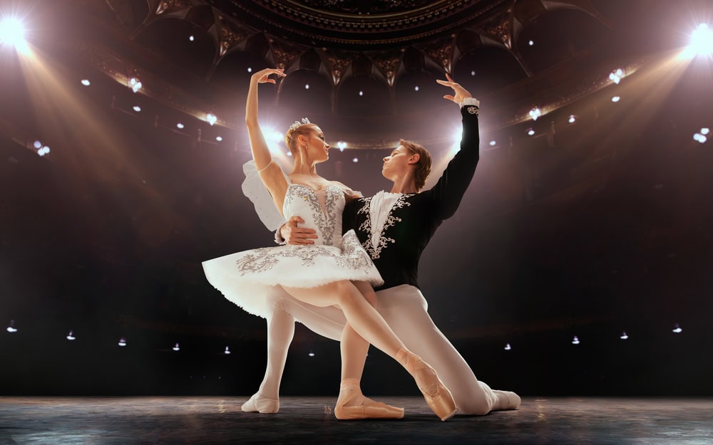 Two ballet dancers perform a pose on stage under spotlights, wearing traditional ballet costumes, with an ornate theater ceiling visible above them.
