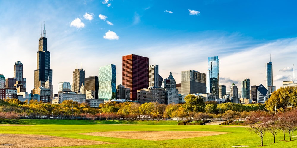 Chicago skyline with tall buildings and skyscrapers in the background, a large green park and baseball field in the foreground, under a blue sky.