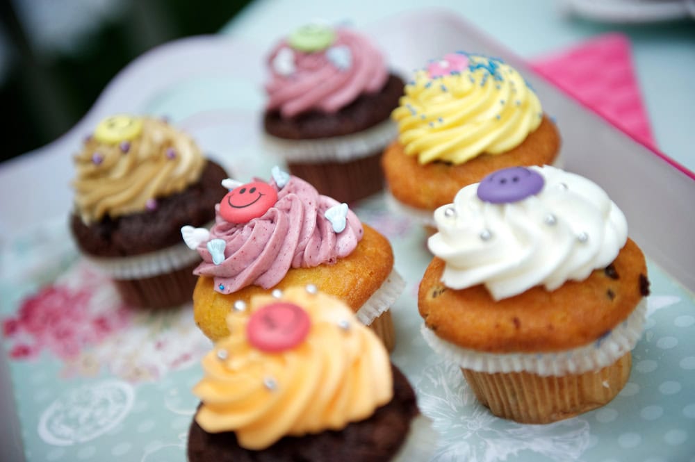 A tray of six decorated cupcakes with colorful frosting, sprinkles, and fondant buttons on top, arranged on a patterned surface.