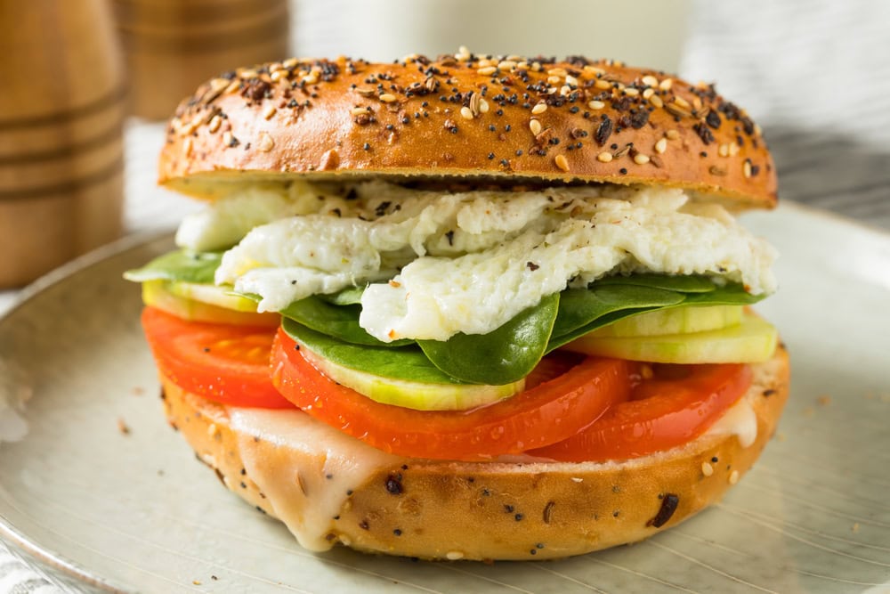 Everything bagel sandwich with egg whites, spinach, cucumber, tomato, and melted cheese on a plate.