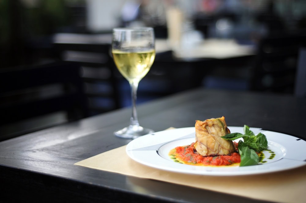A glass of white wine next to a plated gourmet dish with sauce and garnish on a dark table in a restaurant setting.