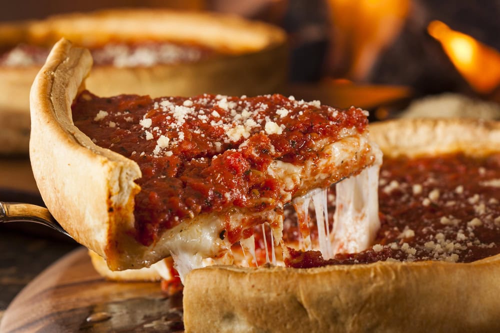 A slice of deep-dish pizza with tomato sauce and melted cheese being lifted from a whole pizza, with a blurred fire in the background.