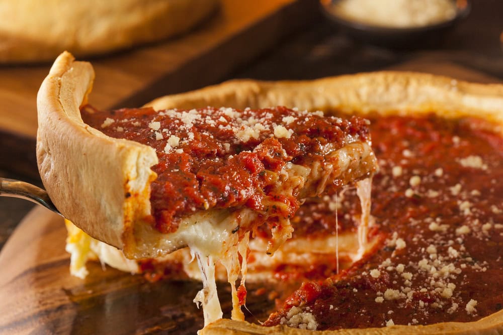 A slice of deep-dish pizza with melted cheese and tomato sauce is being lifted from a whole pizza on a wooden surface.