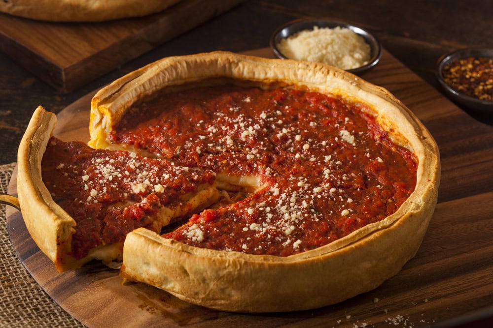 A Chicago-style deep dish pizza with tomato sauce and grated cheese on top, one slice partially removed, served on a wooden board.