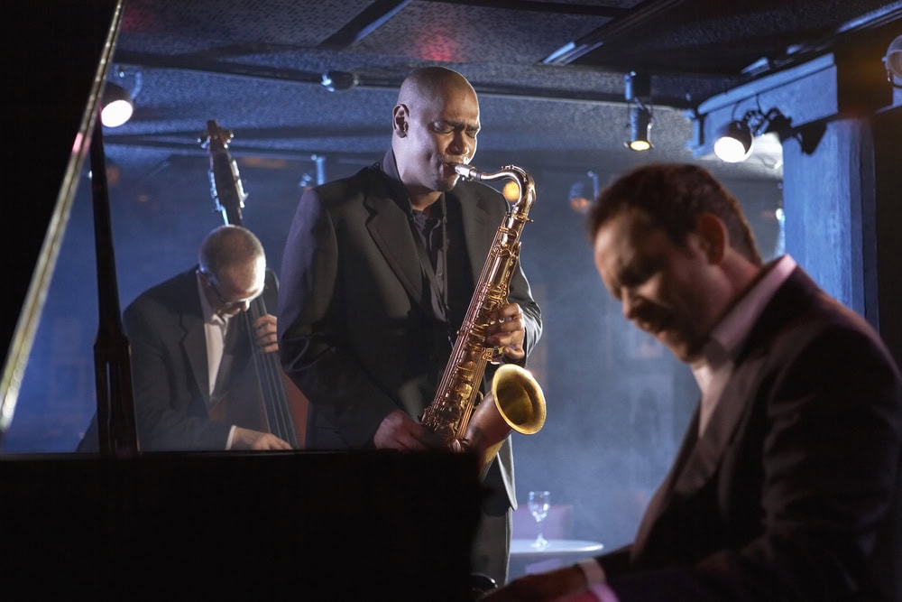 Three musicians perform on stage: a saxophonist plays in the center, a double bassist is in the background, and a pianist is in the foreground. The setting is dimly lit and smoky.