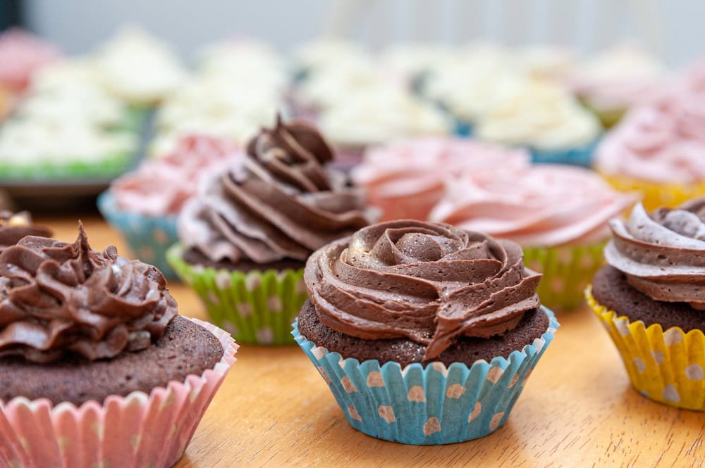 Assorted cupcakes in colorful paper liners with swirled chocolate and pink frosting on a wooden table, with more cupcakes blurred in the background.