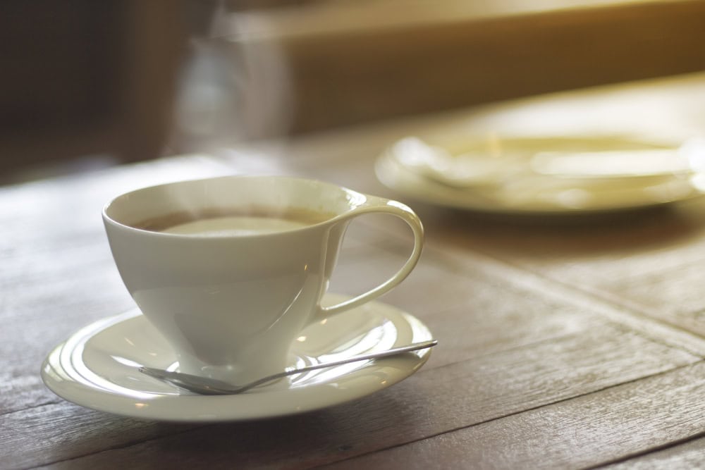 A white cup filled with coffee sits on a matching saucer with a spoon, on a wooden table; a blurred plate is in the background.