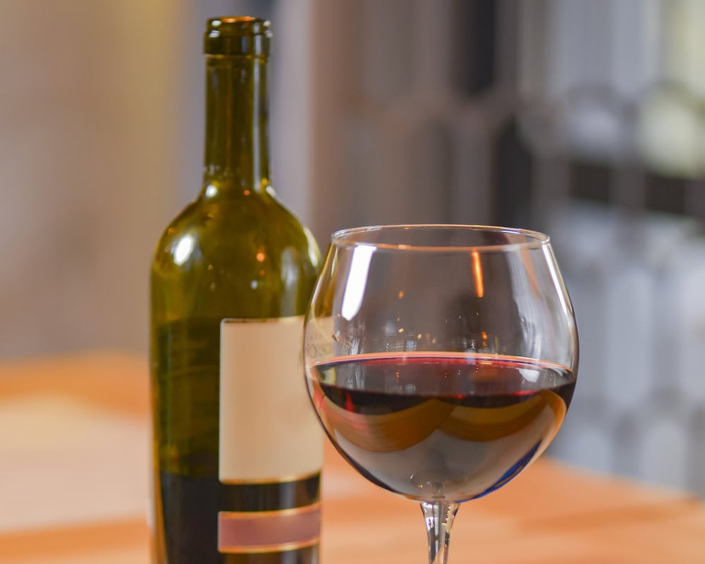A bottle of red wine and a filled wine glass are placed on a wooden table, with a blurred background.