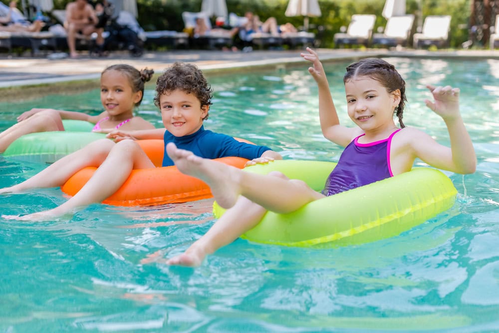 Three children sit in colorful inflatable rings, floating and smiling in a swimming pool on a sunny day.