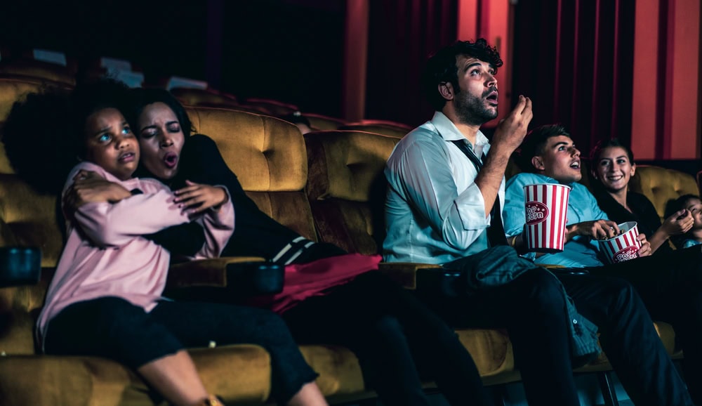 Four people sit in a movie theater, watching a film with expressions of surprise and suspense, holding popcorn and drinks in red seats.