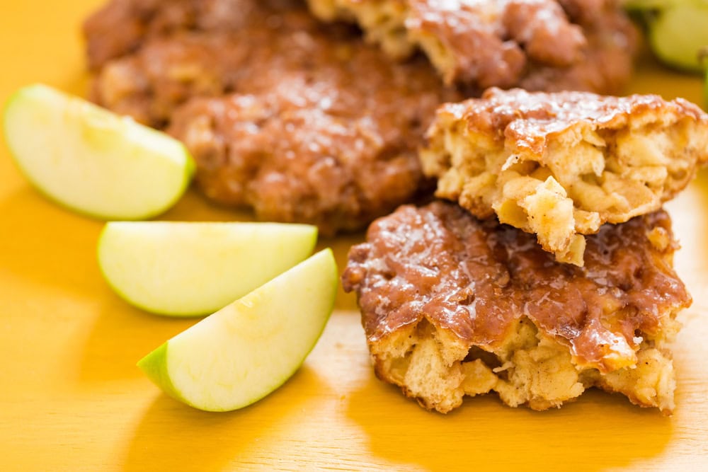 Close-up of apple fritters with a glazed coating next to sliced green apples on a yellow surface.
