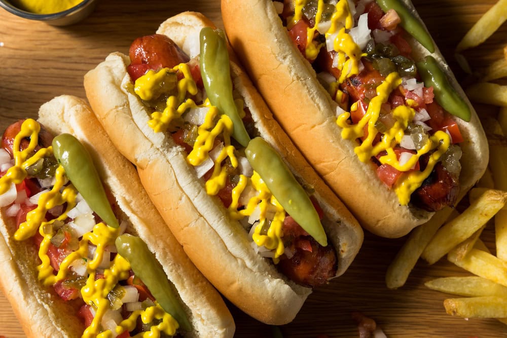 Three Chicago-style hot dogs topped with mustard, onions, relish, tomatoes, peppers, and pickles sit on a wooden surface next to a pile of French fries.