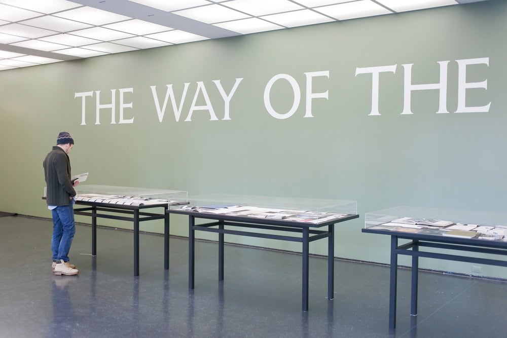 A person stands reading in front of display tables with documents, beneath large text on the wall that reads "THE WAY OF THE.