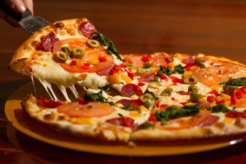 A slice of pizza with cheese, tomato, pepperoni, green olives, red peppers, and herbs being lifted from a whole pizza on a yellow plate.