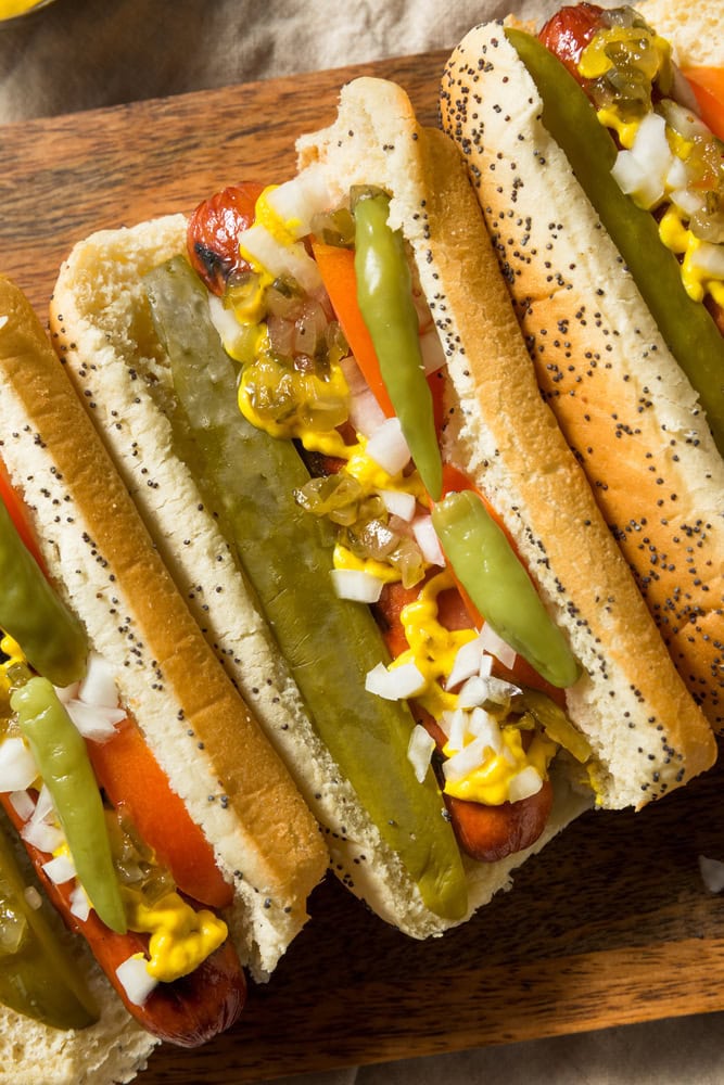 Three Chicago-style hot dogs in poppy seed buns topped with mustard, relish, onions, pickle spears, sport peppers, and chopped tomato, arranged on a wooden board.