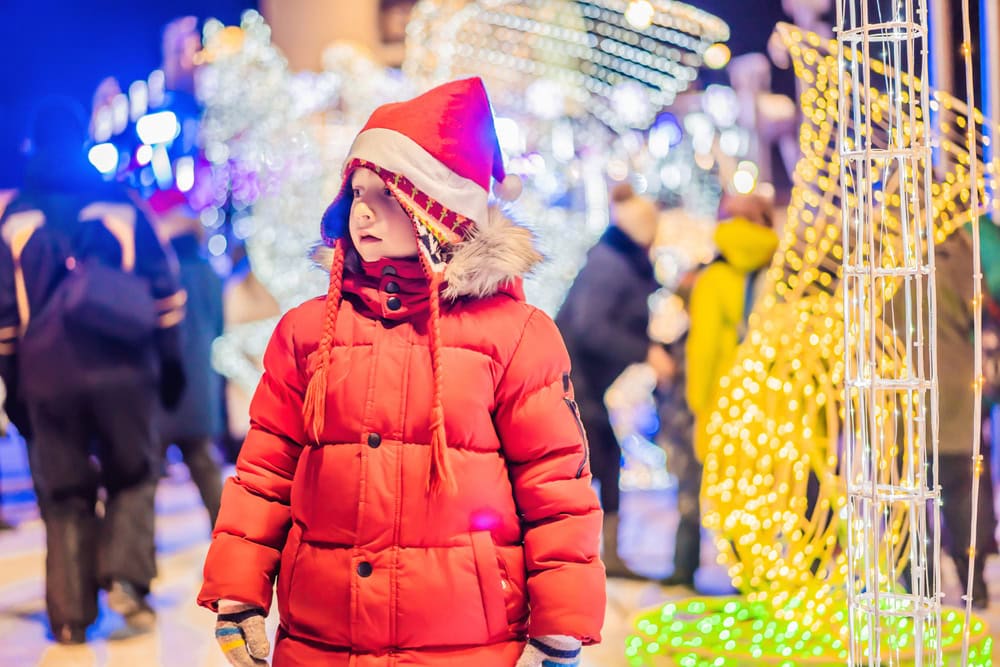 A child in a red winter coat and Santa hat stands outside at night, surrounded by festive holiday lights and people in the background.