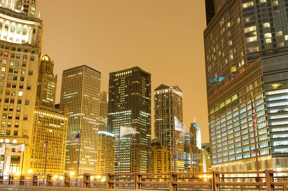 Tall office buildings illuminated at night under a cloudy sky, with reflections visible in the glass facades and a fence in the foreground.