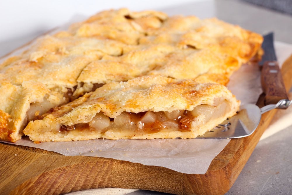 A rectangular apple pie with a golden-brown crust sits on parchment paper atop a wooden board, with one slice partially removed using a metal spatula.