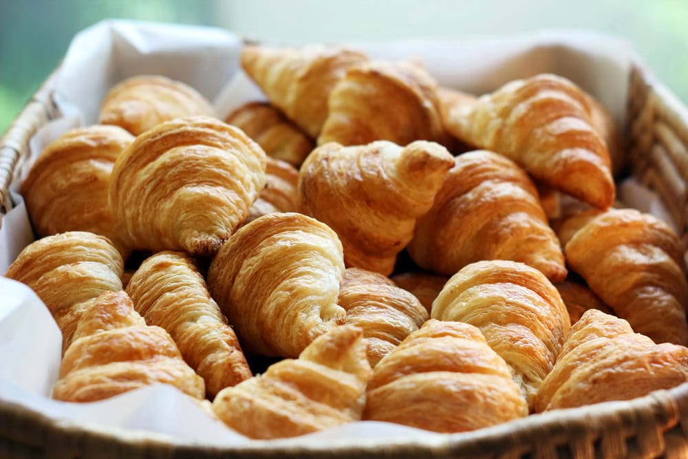 A basket filled with multiple golden-brown croissants, arranged closely together on white parchment paper.