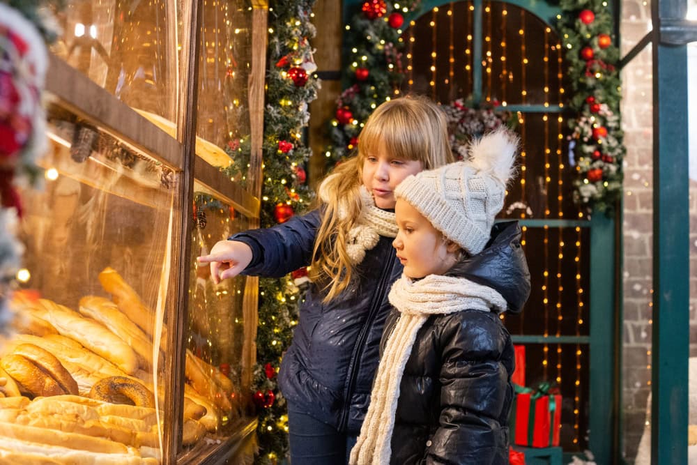 Two children in winter clothing look at a display of bread and pastries at a decorated outdoor market with Christmas lights and festive decorations.