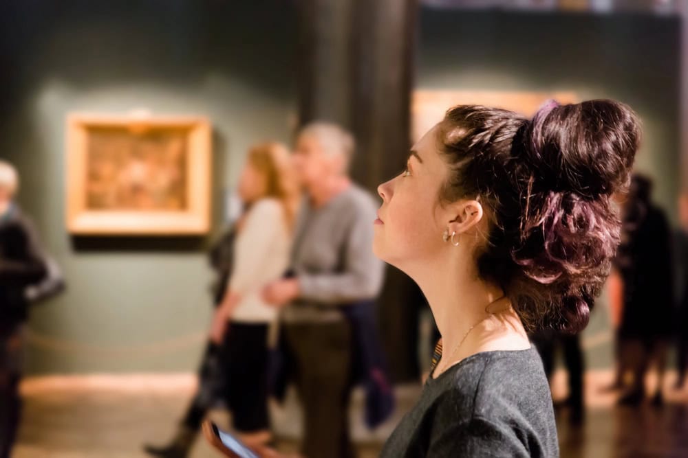 A woman with dark hair in a bun looks at artwork in a museum; blurred visitors and framed paintings are visible in the background.