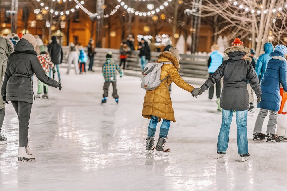 People ice skating outdoors at night under string lights, wearing winter clothes and holding hands on a rink surrounded by trees and festive lighting.