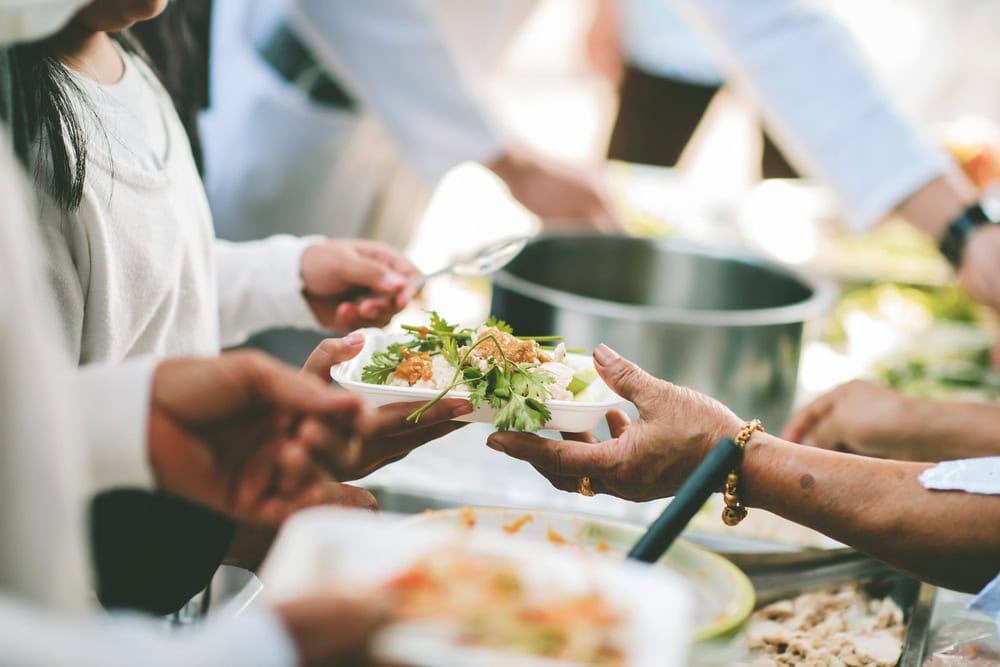 People are serving food onto plates from a buffet-style setup, with hands passing a plate of food topped with greens.