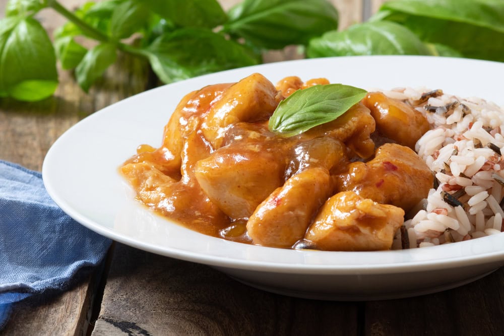 A white plate with chicken curry in sauce garnished with a basil leaf, served alongside a portion of mixed rice; fresh basil leaves in the background.