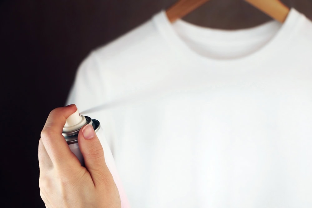 A hand sprays an aerosol can onto a plain white t-shirt hanging on a wooden hanger against a dark background.