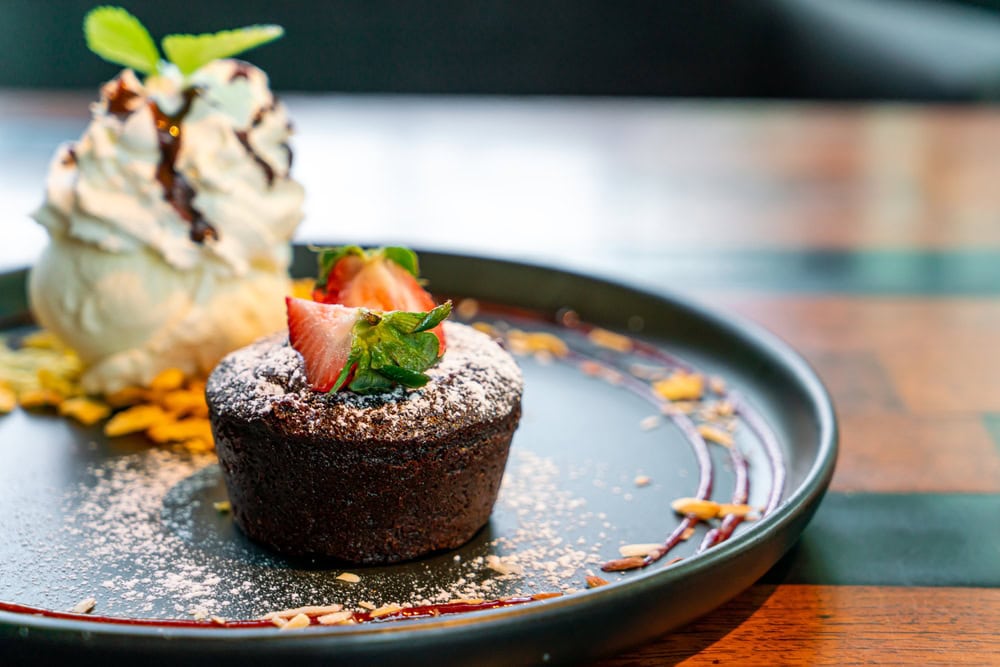 A chocolate lava cake topped with strawberries and powdered sugar is served with a scoop of vanilla ice cream and chocolate drizzle on a black plate.