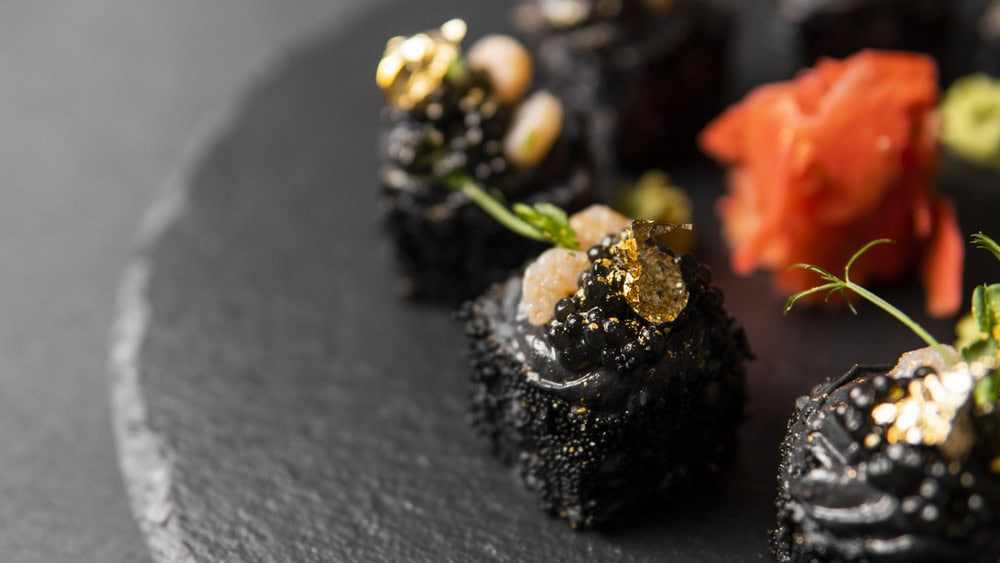 Close-up of gourmet black sushi topped with gold leaf, roe, and garnish, served on a dark plate with pickled ginger and wasabi in the background.