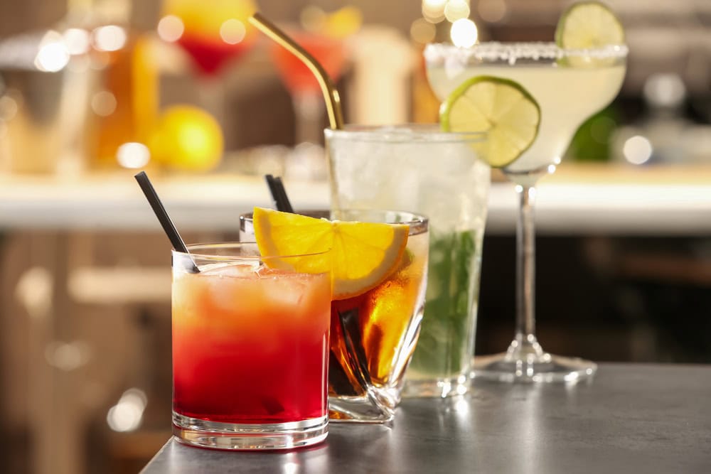 Four assorted cocktails in glasses with garnishes such as orange slices and lime, arranged on a bar counter with a blurred background.