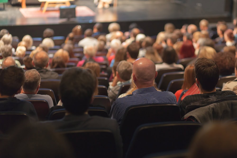 A large audience sits in rows of seats facing a stage, watching an event or performance in a theater or auditorium.