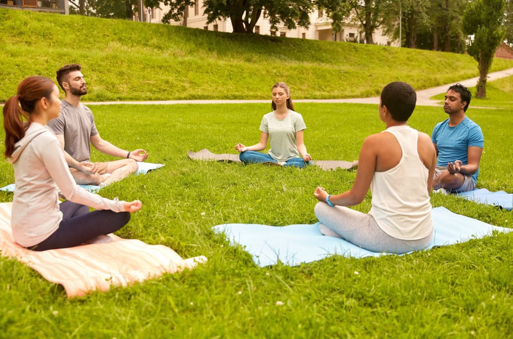 Five people sit cross-legged on yoga mats in a grassy park, eyes closed and meditating in a circle.