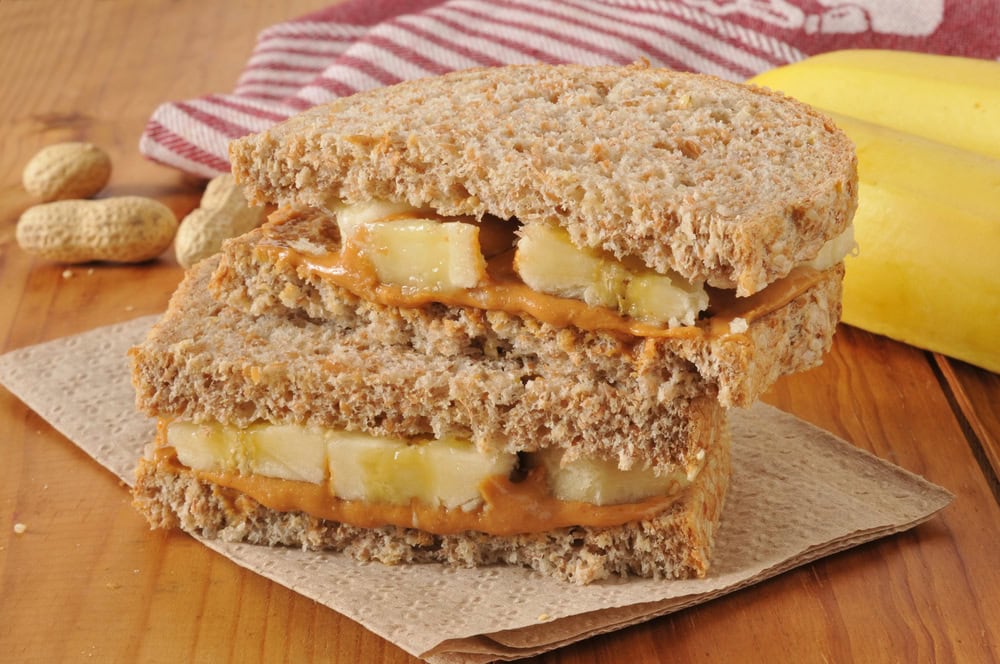 A peanut butter and banana sandwich on whole wheat bread sits on a napkin, with a striped cloth, whole peanuts, and a banana in the background.