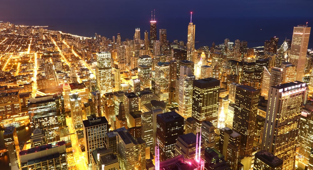 Aerial view of a city skyline at night with illuminated buildings and streets, adjacent to a large body of water.